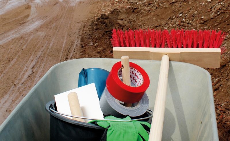 Auf einem Schubkarren sind verschiedene Werkzeuge und Materialien zu sehen: ein Besen mit rotem Kopf, Klebeband in roter und grauer Farbe, eine Rolle Klebefilm und ein grünes Handtuch. Der Schubkarren steht auf einem unebenen, kiesigen Boden.